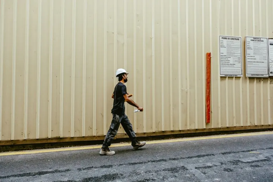 An adult male construction worker walks along an outdoor construction site wall.